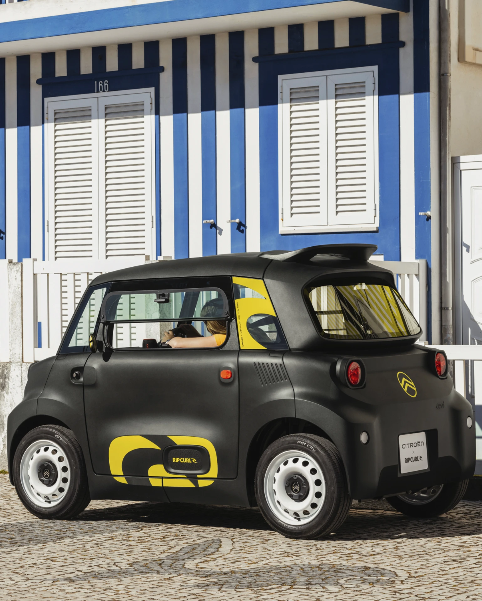 Zij‑achteraanzicht van de Citroën Ami x Rip Curl in een stadsstraat, elektrische auto zonder autorijbewijs, met extra stabiliteit dankzij vier wielen.