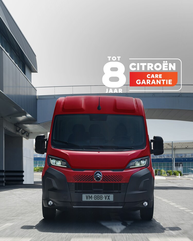 Voorzijde van een rode Citroën Jumper die door een moderne stedelijke omgeving rijdt, waarbij het robuuste ontwerp, het praktische karakter en de krachtige uitstraling worden benadrukt.